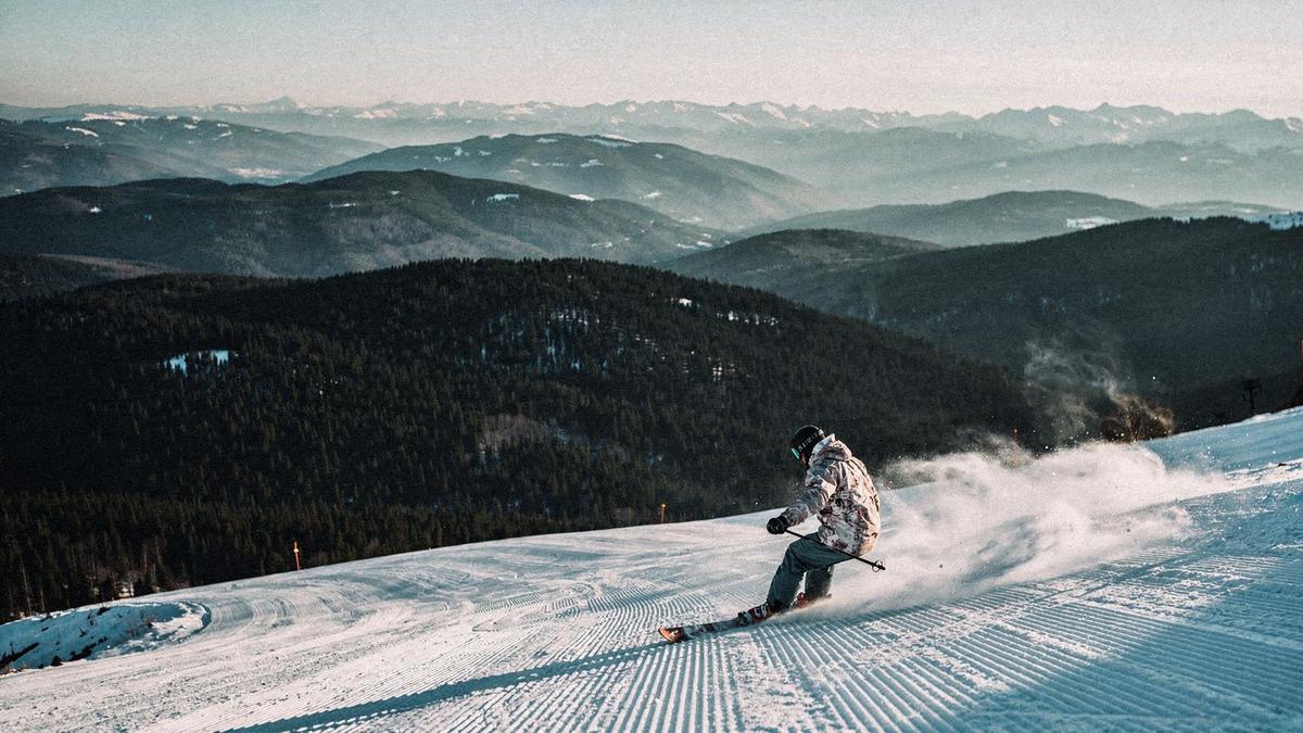 Skier carving a groomed piste above a pine valley in late afternoon light