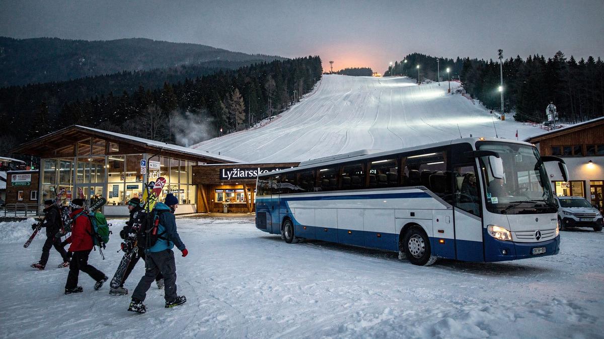 Coach waiting at the base station of a snow-covered mountain resort at dusk while skiers carry gear toward the lift